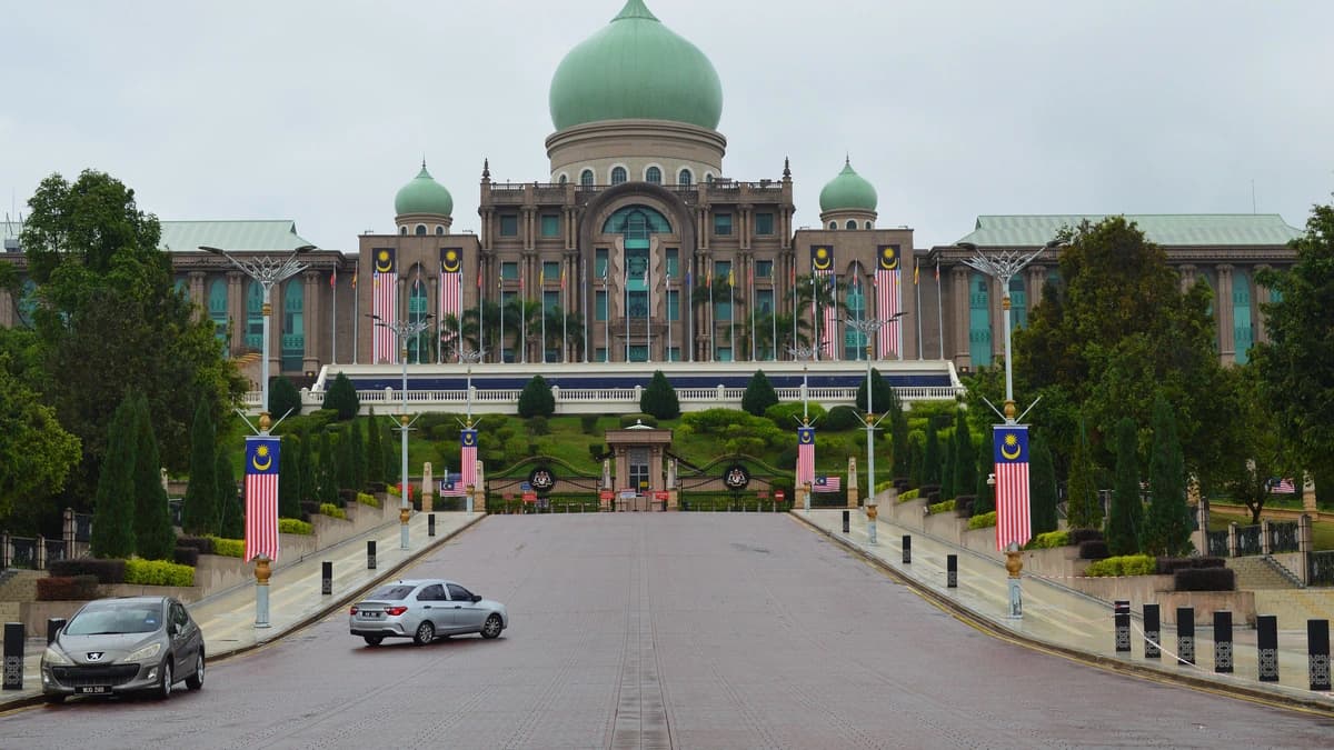 Green-domed Perdana Putra building in Putrajaya at the end of a wide road, with Malaysian flags lining the approach under an overcast sky