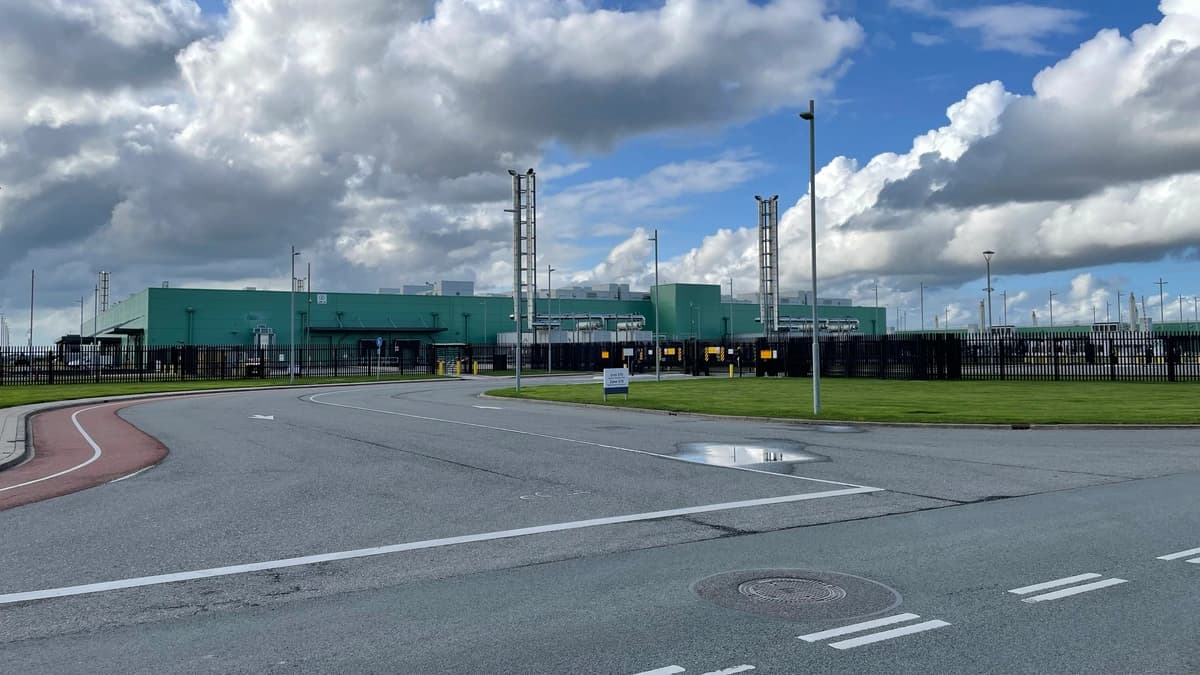 Green industrial data centre building behind a fence and grass verge under a blue sky with large clouds, seen from a road junction