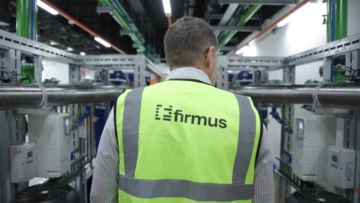 Worker in a high-visibility Firmus vest walking between rows of cooling and electrical plant inside an AI factory