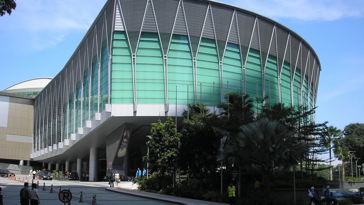 Curved glass and steel facade of the Kuala Lumpur Convention Centre above a street entrance, with palm trees and a few pedestrians