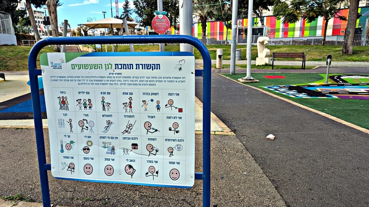 Blue-framed board in a public park showing rows of simple drawn symbols with Hebrew labels, beside a green playground surface