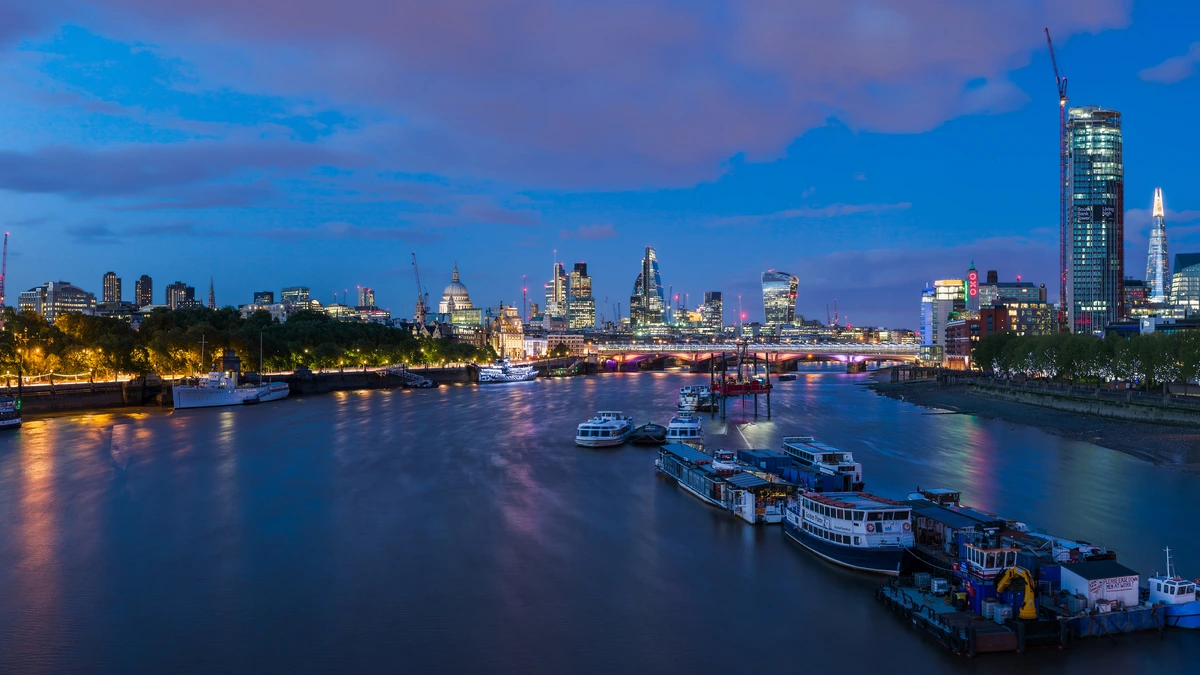 The City of London skyline at dusk seen across the Thames, with boats moored in the foreground and a domed cathedral at centre