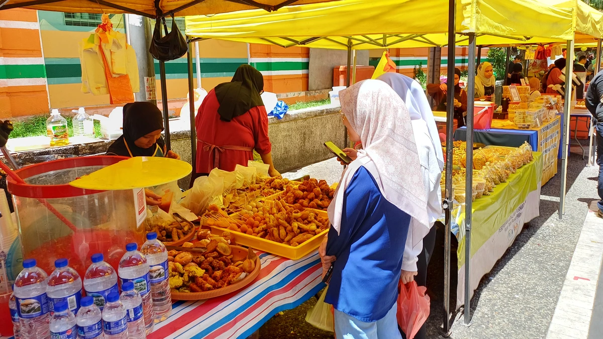 A woman in a pink headscarf looks at her phone while standing at a street food stall under yellow canopies at a market on Jalan Benteng, Kuala Lumpur, with trays of fried snacks and bottled water on the counter