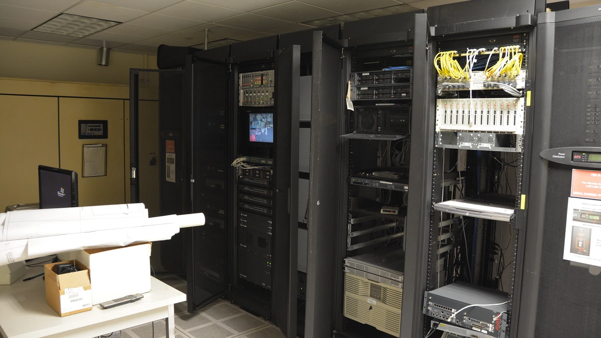 Dark server racks with open doors in a small server room, yellow network cables in one cabinet and a desk with a monitor and rolled plans beside them