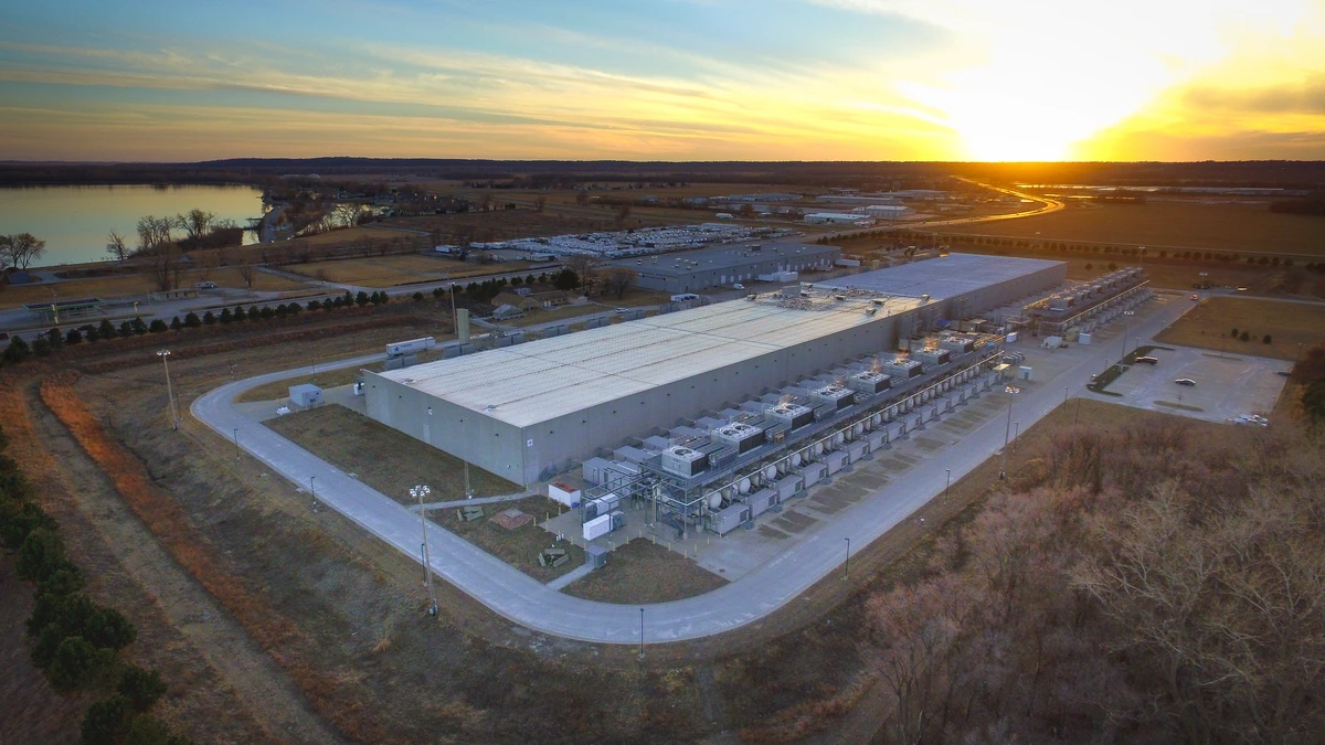 Aerial view at sunset of a long grey data-centre hall with rooftop cooling equipment, a looping access road and a lake on the horizon