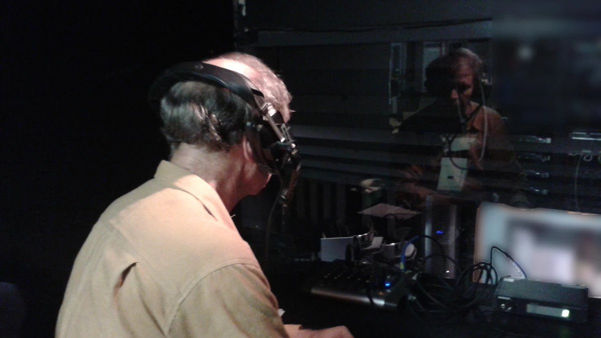 A man wearing headphones sits in a darkened booth in front of mixing equipment, his reflection visible in the glass