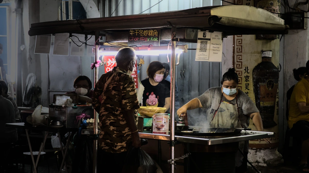 A hawker in a face mask cooking char koay kak in a large wok at a night stall in George Town, Penang, while masked customers wait under a fluorescent light
