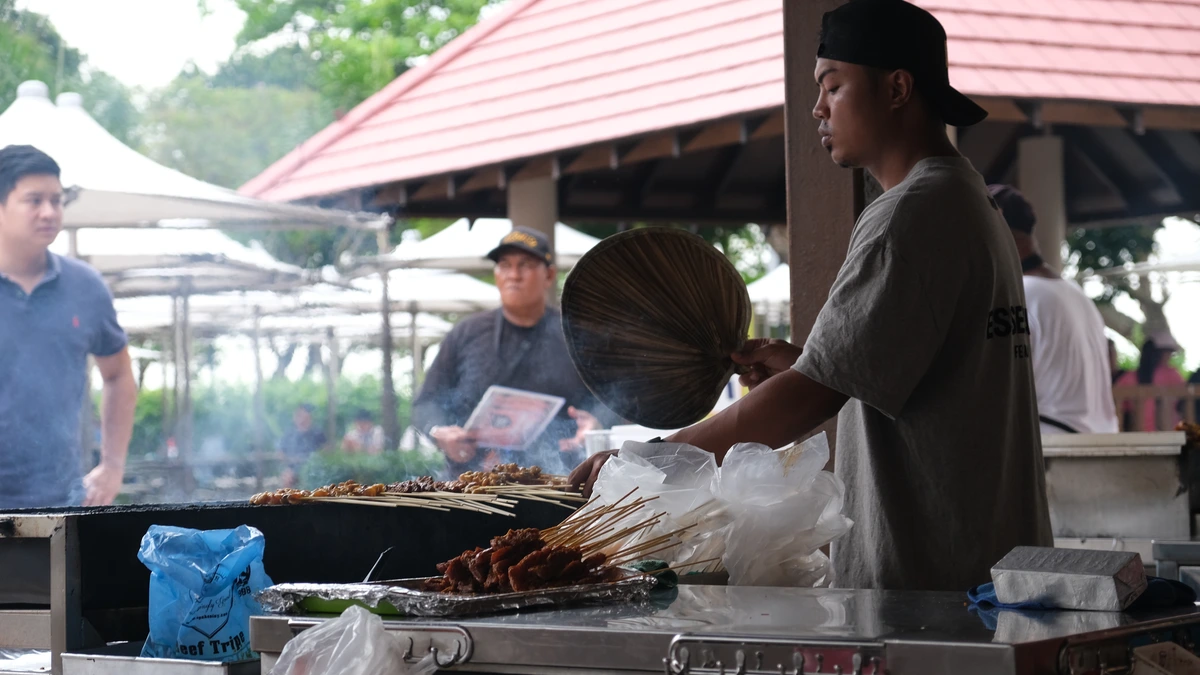 A satay seller in a black cap fanning skewers of grilled meat with a woven fan at East Coast Lagoon Food Village in Singapore, while customers wait in the background