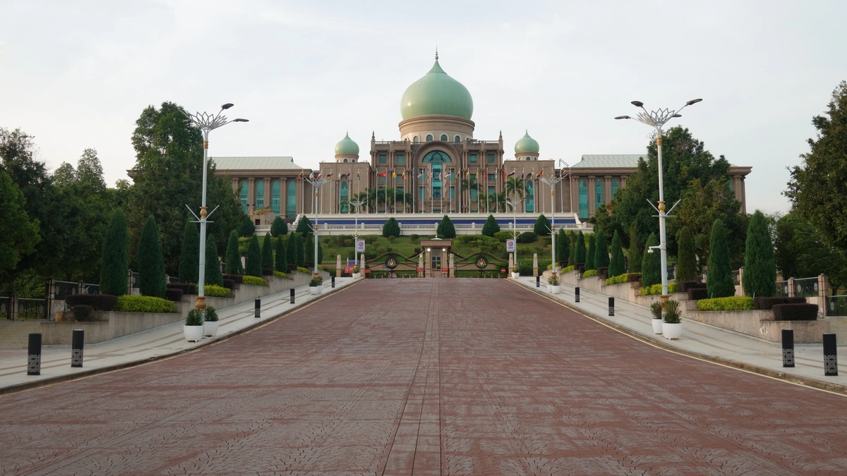 A long red-brick boulevard lined with street lamps and cypress trees leads to a large building with a green central dome and rows of flags