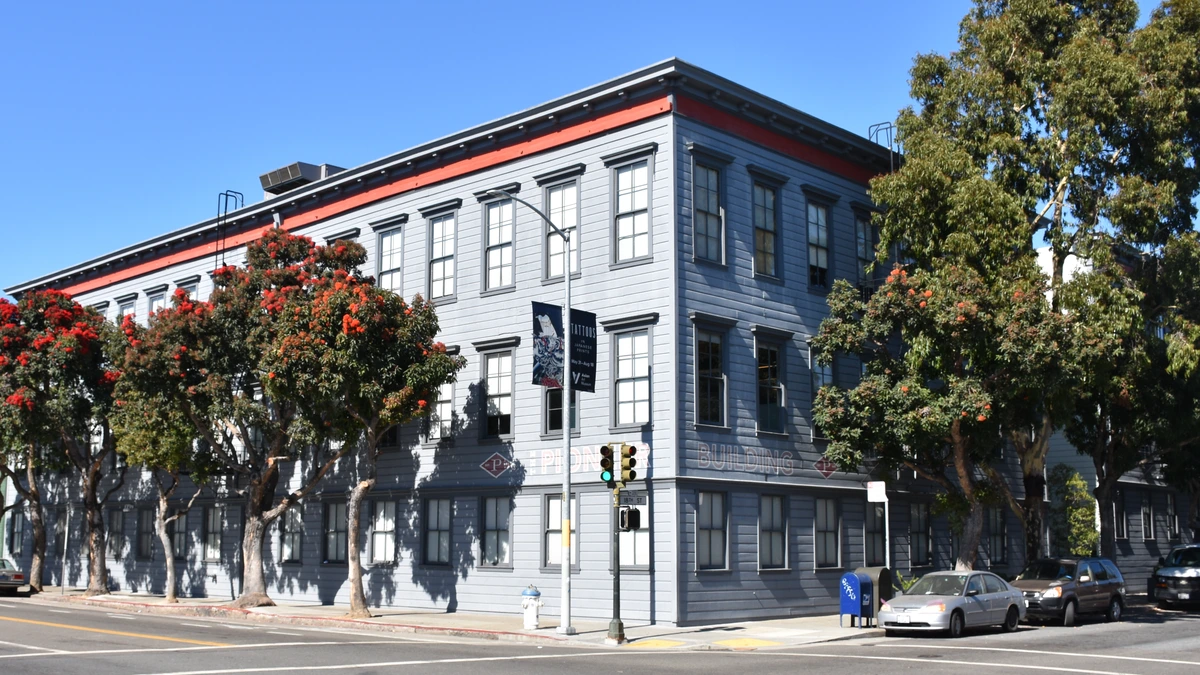 A three-storey grey wooden building with red trim on a San Francisco street corner, with trees in red bloom and cars parked in front