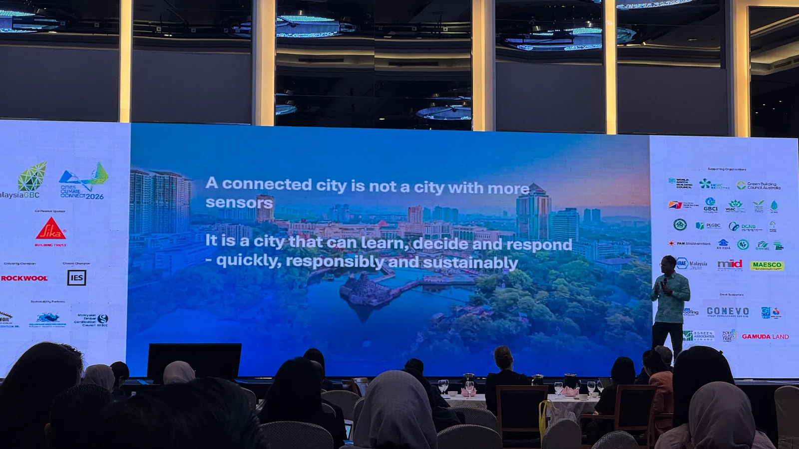 A speaker addresses a seated conference audience beside a wide screen reading that a connected city is not a city with more sensors