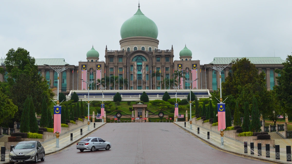 A green-domed government building at the end of a wide road lined with Malaysian flags under an overcast sky